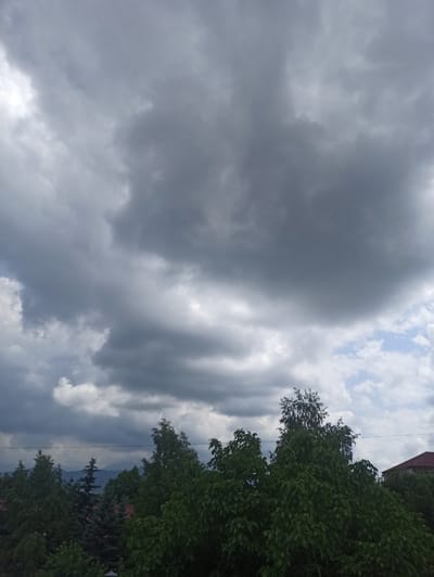 The image captures a dramatic sky with a mix of dark, heavy clouds and patches of lighter sky with some white clouds. The sky takes up a significant portion of the frame, suggesting an impending weather change or recent storm. To the right, lush green trees with dense foliage are visible, indicating a natural or semi-natural setting. A hint of a red-tiled roof can be seen peeking from behind the trees, suggesting the presence of buildings nearby. The overall impression is one of a slightly overcast or stormy day, possibly late afternoon or early evening, given the contrast between the dark clouds and the illuminated sky. The location context of Dariali, Georgia, implies a mountainous or valley region, which could be further supported by the distant dark shapes that might be mountains. There is no activity or event clearly depicted, and no visible text. The emotions evoked are a sense of the power of nature and the changing moods of the sky.