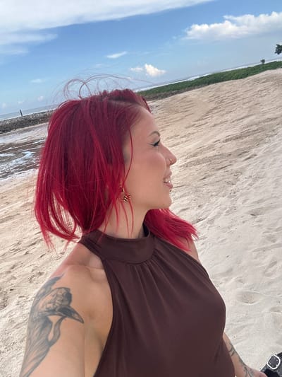 A woman with red hair and a brown dress is outdoors, seemingly on a beach, in Nusa Dua, Indonesia. She has a tattoo on her arm and is smiling and looking up towards the sky. The environment includes a sandy beach, the ocean, and the sky with clouds.