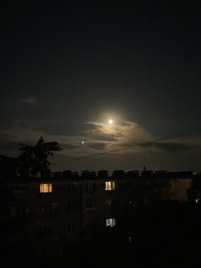 This is a nighttime photograph of an apartment building in Yevpatoriya, Ukraine, under a cloudy sky with a bright moon. The moon is partially obscured by clouds, creating a halo effect. There is a faint greenish object visible in the sky to the left of the moon, possibly a satellite or a planet.

The apartment building is dark, with only a few windows illuminated. One window on the left side of the building emits a warm, orange light, suggesting an interior light source. Another window on the right side of the building also shows a faint light. The building's facade is not clearly visible due to the darkness.

In the foreground, to the left of the building, a silhouetted tree with dense foliage is visible. Power lines run horizontally across the upper portion of the building. The overall atmosphere of the image is quiet and serene, with a sense of urban night.
