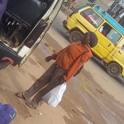 A woman stands on a street in what appears to be Sardauna, Nigeria. She wears an orange jacket, brown pants, and sandals. She carries a white plastic bag. Behind her is a yellow and blue vehicle with the word "Turbo" visible on the side. To the left, partially obscured, is another vehicle. The street surface appears to be a combination of dirt and paved areas, with some debris scattered around. The environment suggests an urban setting.