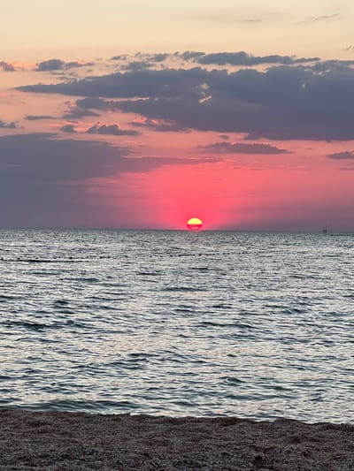 The image depicts a sunset over the sea. The sun, a vibrant orange orb, is partially submerged on the horizon, casting a warm glow across the sky and water. The sky is a gradient of soft pinks and oranges, transitioning into a deep blue with scattered clouds. The sea is a rippling expanse of blues and greys, reflecting the colors of the sky. A dark, textured foreground, likely a beach or shoreline, occupies the right side of the frame. The overall mood is serene and tranquil, suggesting a peaceful end to the day. The image is oriented vertically, giving a sense of depth and expansiveness to the seascape. There is no visible text in the image.