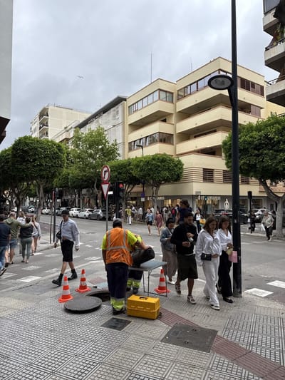 This image depicts an active urban street scene in Eivissa, Spain, specifically along Avinguda d'Espanya, with the "EMPRESAS MALUTES" building visible at number 12 and the "ST. HILARIUS" shop at number 18. The sky is overcast, indicating a cloudy daytime.
In the foreground, a public utility crew is performing maintenance on an open manhole on the patterned tiled sidewalk. Three orange traffic cones surround the manhole, with its circular cover placed alongside. A yellow portable toolbox rests next to the manhole. Two workers are visible; one, wearing an orange high-visibility vest and dark trousers, is bent over the manhole with his back partially to the viewer. Another worker, also in work attire, is partially obscured, assisting with a black, rolled-up object placed on a small grey metal table positioned nearby.
Numerous pedestrians navigate the sidewalk and street. A man in a light-colored collared shirt, dark shorts, and black boots walks directly past the roadworks, looking towards the camera. Further into the midground, a woman in a white long-sleeved top and white pants walks towards the camera, carrying a small handbag. Beside her, a man in a dark top and shorts holds a white cup. To their left, another woman in a light-colored dress observes the roadworks. Various other individuals are scattered across the sidewalk and street, some walking, others in front of shop windows. A man carries a child on his shoulders further down the street. In the far background, a person is seen running across the street.
The street is lined with numerous small, spherical-canopy trees. Parked cars are visible along the street in the background. Buildings rise on both sides; on the left, a light-colored multi-story building prominently features the "EMPRESAS MALUTES" sign. Adjacent to it, a yellowish/cream building with multiple balconies and distinct horizontal facade elements is visible. Shopfronts, including "ST. HILARIUS," line the base of the buildings. A black lamppost stands in the far-right foreground. A traffic light showing a red signal for vehicles and a red circular "no right turn" sign are visible near the roadworks.