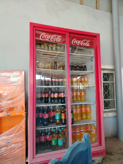 In the city of Usto Xo‘jamat, Uzbekistan, an indoor scene captures a commercial establishment's beverage display. The focal point is a red-framed glass-door refrigerator, filled with an assortment of bottled drinks. On the left, a stacked display of orange Fanta bottles, wrapped in plastic, stands beside the refrigerator.
Inside the refrigerator, the left section displays several shelves. The top shelf appears to hold an assortment of smaller bottles or cans, some with yellow labels. The shelves below are stocked with numerous dark-colored bottles, likely cola beverages, with red and white branding visible. Mixed with these are some bottles of a lighter colored beverage, possibly Fanta or a similar orange-flavored drink, identifiable by their orange liquid and branding.
The right section of the refrigerator mirrors the organization of the left, with a variety of drinks. The top shelves contain various bottles, including what appear to be darker sodas. Below these, several shelves are filled with orange-colored bottled drinks, clearly labeled as Fanta. The bottom shelves also show a continuation of these orange-colored bottles. A clear plastic fan is visible in the upper left portion of the right section.
The environment surrounding the refrigerator consists of plain, light-colored walls. To the right of the refrigerator, a window with a distinctive criss-cross pattern in its panes is present. The floor appears to be concrete. The lighting suggests an indoor setting, likely a shop or convenience store. A portion of a blue object, possibly a chair or a piece of furniture, is in the foreground, partially obscuring the bottom of the refrigerator. The overall impression is one of a well-stocked beverage retail space.