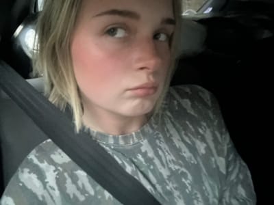 A woman with blond hair is in a car, wearing a gray camouflage shirt and a seatbelt. The location is not discernible in the image.