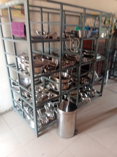 The scene is an interior view within Ejule, Nigeria, depicting a storage area for kitchen or catering equipment. A large, gray, multi-tiered metal shelving unit, approximately 2 meters high and 3 meters wide, dominates the image, extending from the left to the right side of the frame. This unit holds numerous items of reflective, clean stainless steel cookware and serving ware, including various sizes of pots, pans, bowls, platters, and ladles, systematically arranged on its multiple shelves. A folded purple towel is visible on the top left shelf. On the upper right shelves, several plastic containers and coiled hoses or cables are present. A cylindrical, stainless steel pedal-operated waste bin with a domed lid stands on the light-colored tiled floor in front of the lower right section of the shelving unit. The background wall is light-colored. No individuals are present, and no actions are observed.