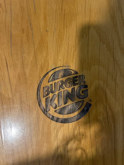 The image displays a dark gray Burger King logo, featuring the wordmark "BURGER KING" enclosed within two bun-like semicircles, etched or printed onto a light brown wooden surface. The wood exhibits prominent vertical grain lines, numerous fine scratches, and scattered specks of dust. The letters in "BURGER KING" are capitalized, bold, and contain a hatched texture within them, giving the appearance of depth. A small registered trademark symbol (®) is visible to the right of the word "KING." The wooden surface shows varied lighting conditions, with a distinct brighter, more reflective area in the lower left portion of the frame, contrasting with a darker, shadowed region that dominates the right side.