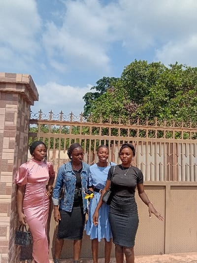 Four young adult females are standing outdoors in Ejule, Nigeria, in front of a brown metal gate with decorative finials. To the immediate left is a tiled structural pillar. From left to right, the first female wears a ruched pink gown and holds a black handbag. The second wears a denim jacket over a black top and skirt, with a yellow item on her hip. The third wears a light blue dress. The fourth wears a black top and skirt with a black shoulder bag, her left hand outstretched with an open palm. Behind the gate, dense green foliage is visible. The sky above is blue with scattered white clouds, indicating bright daylight. The ground beneath the individuals is an unpaved, light brown surface.