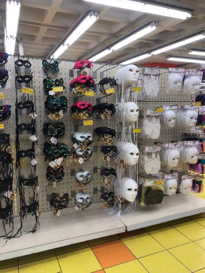 An indoor, overhead shot captures a vibrant display of masks and accessories for sale on a white pegboard wall, likely in a craft or party supply store in Marousi, Greece. The store is brightly illuminated by multiple rows of fluorescent light fixtures running along the left side of the frame.
The display features various types of masks. Several rows of plain white, full-face masks are visible, some individually packaged in clear plastic. Below these are rows of white paper masks, some with printed designs, also in clear packaging. Further down, there are numerous half-face masquerade-style masks. These come in a variety of styles and colors, including sparkly pink, silver, black, and iridescent sequined designs. Many of these smaller masks are packaged with small yellow tags that likely indicate prices, with some appearing to show euro symbols and numerical values, such as "€3.50" or "€4.00." Barcodes are also visible on some tags.
The pegboard is densely packed with hooks holding the merchandise. To the right, a distinctive wall with large yellow and orange square tiles is visible, providing a contrasting background to the mask display. No people are visible, suggesting the image captures a static product arrangement during store hours. The bright artificial lighting indicates an indoor setting, likely during the day or evening.