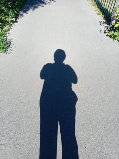 The image shows the shadow of a person standing on an asphalt path. The path is bordered by green vegetation on the left and a colorful fence and more vegetation on the right. The sun is casting a long shadow, indicating it is likely morning or late afternoon. The scene appears to be a park or a walking trail in an outdoor setting. The city of Novotroitsk, Russia, is mentioned as the location, suggesting the path is within or near the city. There are no other people or objects clearly visible in the shadow or the immediate surroundings.