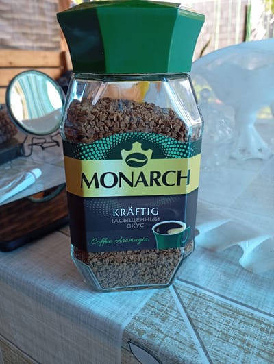 This media file features a glass jar of Monarch brand instant coffee. The jar is hexagonal and filled with dark brown coffee granules. The label is primarily black with a yellow band across the middle. The word "MONARCH" is prominently displayed in large, bold, black letters on the yellow band, with a green stylized crown and coffee bean logo above it. Below the brand name, the label contains text in both German and Russian. The German text reads "KRÄFTIG," and the Russian text below it translates to "насыщенный вкус" (rich taste). Further down, in green script, is "Coffee Aromagia." A small illustration of a green coffee cup with white foam sits to the right of the text. The jar is placed on a table with a light-colored, textured surface, possibly made of wood or a similar material, with a grid pattern. In the background, a white, sheer fabric is visible to the right, and a wooden structure and a round mirror are partially visible to the left. The lighting suggests it is daytime, possibly outdoors or in a well-lit indoor space. The overall scene is casual and appears to be set in a domestic or outdoor seating area. There are no people visible in the image. The presence of a tube of what appears to be toothpaste and a smartphone on the table hints at a personal setting. The location context provided is Urus-Martan, Russia.
