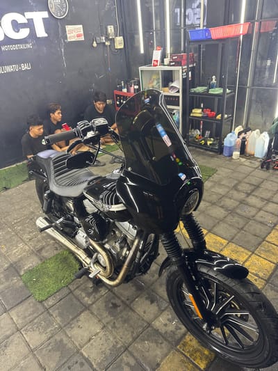 Here is a detailed description of the scene:
The image shows a black motorcycle parked indoors. The location is a "MOTODETAILING" shop, specifically "OCT MOTODETAILING ULUWATU - BALI" indicated by a sign on the wall. Uluwatu is located in Ungasan, Indonesia.
Several men are present in the shot; they seem to be examining or detailing the motorcycle. The motorcycle has a sleek design, and its front wheel is turned slightly to the right. The floor is tiled, and there are patches of artificial grass surrounding the motorcycle. Shelves filled with cleaning supplies are visible in the background. The shop appears well-lit and organized.
