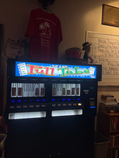 The image shows an indoor scene, likely within a community center or club, depicting a "Full Tabs" gaming machine as the central focus. The machine has a bright, illuminated display that reads "PLAY & WIN Full Tabs" with cartoonish graphics of game symbols and a logo that says "AMERICAN GAMES." Below the display are rows of slots where game tickets are inserted or dispensed, with some tickets already visible. The machine has multiple "PLAY" buttons and digital readouts displaying numbers, suggesting it is operational and in use.
To the left of the gaming machine, an American flag, partially obscured, is visible. Above the machine, a red t-shirt hangs, displaying text that reads "American Legion Post 316," "Atlantic Beach, FL," and "REMEMBER EVERYONE DEPLOYED," indicating a connection to the American Legion Post in Atlantic Beach, Florida.
To the right of the gaming machine, a whiteboard with handwritten notes lists events such as "NASCAR DISCOUNT DAY," "BINGO," and "STEAK OR SAL." Below the whiteboard, a bookshelf is filled with various books, and a trash can is partially visible. The overall environment appears somewhat dimly lit, with the gaming machine's illumination being the primary light source in the foreground. The background suggests a casual, communal space, possibly a hall or recreation room associated with the American Legion post.