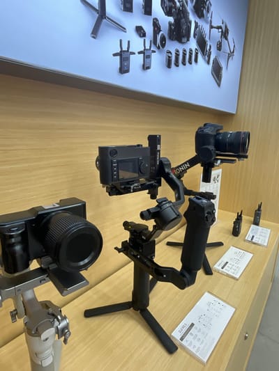 The image depicts a retail display of camera equipment in a store, likely in Wichit, Thailand, given the context. In the foreground, a silver camera gimbal is visible on the left, topped with a black camera and lens. To its right, a black DJI Ronin gimbal is prominently displayed, holding a black camera with a lens. Several small black devices, possibly wireless transmitters, are placed on the wooden counter surface to the right of the Ronin. Two informational pamphlets, one labeled "DJI RS3" and another with similar branding, are laid out on the counter. In the background, a large light blue display screen showcases various DJI products, including drones and camera accessories, mounted on a white background. The overall environment suggests a modern electronics store. There are no people visible in the image.