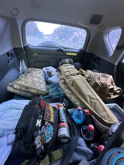 The image shows the interior of a vehicle, likely a SUV, with the rear cargo area filled with various items. The view is from inside the vehicle looking towards the rear window, which displays a road with trees and buildings visible outside. Several bags, including camouflage-patterned ones, are packed tightly together. A long, tan rifle case lies on top of other items. There are also water bottles and a can of spray. The overall impression is one of prepared travel or relocation, with a substantial amount of gear. The reference to "Verkholy, Ukraine" is not directly evident from the visual information in the image itself.