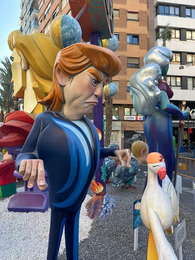 This is a vertical, eye-level, medium shot of a large, colorful artistic installation outdoors. The sculpture features stylized figures with exaggerated features and bright colors. In the foreground, a large, cartoonish figure with ginger hair, a blue jacket with blue stripes, and tan skin is prominently displayed. The figure's left hand is extended, holding a purple object. Behind this figure, a large yellow shape representing hair or clothing is visible.
In the mid-ground, other sculpted elements are present. One is a large, white bird-like figure with a pink beak and cartoonish eyes, perched on a blue body. Further back, a silver-colored abstract sculpture resembles a face or headdress. Other smaller, spherical blue sculptures with gold accents are also visible. The figures are set against a backdrop of a building with a brick facade and white upper floors, featuring numerous windows.
The scene appears to be an outdoor event, possibly a festival or exhibition. The ground is covered in small white pebbles. The lighting suggests it is daytime, with bright sunlight casting shadows. The overall impression is one of vibrant art and festivity. There are no explicit time-of-day or weather cues beyond the general brightness. The location is implied to be Alicante, Spain, as indicated by the context. A small sign with text is visible in the upper right corner, but the text is too small to read. Another sign with text is visible on the white bird-like sculpture. The installation appears to be a large-scale piece of public art, possibly part of a larger display.