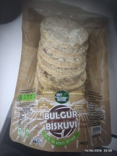 A close-up, overhead shot shows a package of flat, round, light brown, puffed grain crackers. The packaging is primarily brown paper with some translucent plastic. The crackers are stacked in two columns. Text on the packaging is in Turkish and English, with the brand name "UNITY NATURE" prominent. The text mentions nutritional information, ingredients like "BULGUR" (bulgur wheat), and claims of being "PROTEIN KAYNAGI" (protein source). A small barcode is visible, along with a date and time stamp "16/06/2026 20:50" in the bottom right corner. The background is blurred, suggesting an indoor setting with diffuse lighting. There are no people or animals in the image. The location is not explicitly stated in the image, but the text "Türkiye" (Turkey) is present, indicating the country of origin.