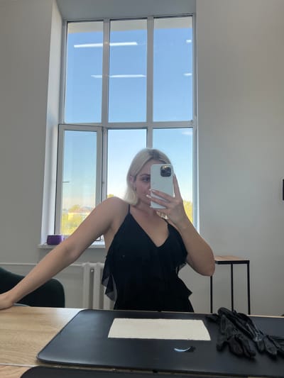 A young woman with blonde hair is taking a selfie in a room with large windows. She is wearing a black dress and holding a white smartphone in her right hand. The room appears to be modern, with a clean, white wall and a wooden desk. Outside the windows, the sky is bright blue, suggesting it is daytime, and there are trees visible in the distance, indicating an outdoor environment. The woman's expression is neutral as she looks at the phone's screen. On the desk, there is a black object that looks like a pair of gloves and a white paper. The overall impression is a casual, everyday moment captured in a well-lit setting.