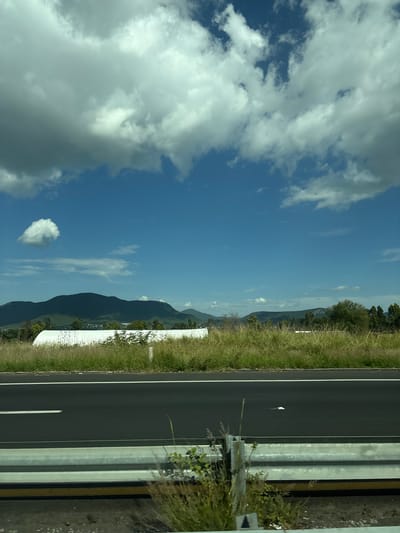 This media file captures a scene from a roadside in Santa Cruz Tehuixpango, Mexico. The perspective is from a moving vehicle, as indicated by the slightly blurred foreground and the road ahead.
The primary subjects are the road, a grassy embankment, and a mountainous landscape under a partly cloudy sky. The road is asphalt with white lane markings, suggesting it is a highway or a significant thoroughfare. A metal guardrail runs alongside the road, separating it from the embankment. The embankment is covered in tall, dry grass, interspersed with some greenery and what appear to be white roadside markers or barriers. In the distance, rolling hills and mountains are visible, covered in a mix of vegetation. The sky is a bright blue with scattered white clouds, some of which are fluffy and prominent, while others are more wispy.
The activity taking place is implied travel along the road. The time of day appears to be daytime, likely late morning or early afternoon, given the brightness of the sky and the shadows. The weather seems pleasant, with clear skies and sunshine, though the clouds suggest it's not completely cloudless.
Notable details include a small, white object on the road, possibly litter or debris, and a single, heart-shaped cloud in the sky. The mountainous terrain suggests a rural or semi-rural location. No people are visible in the frame. No text is discernible in the image.
