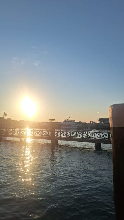 The image depicts a serene waterfront scene at sunset, likely in Nantucket, United States of America, given the architectural style of the buildings and the presence of a ferry labeled "SeaStreak." In the foreground, the calm water reflects the warm, golden light of the setting sun, creating a shimmering effect. A wooden pier with a decorative railing extends across the scene, supported by posts. Beyond the pier, a large white ferryboat is docked, its name "SeaStreak" visible. Several smaller boats are also present in the harbor. In the background, buildings with characteristic New England architecture line the shore, and a flag is visible flying from a building on the left. The sky is a clear, pale blue, transitioning to a warmer hue near the horizon where the sun is setting. The overall atmosphere is peaceful and picturesque.