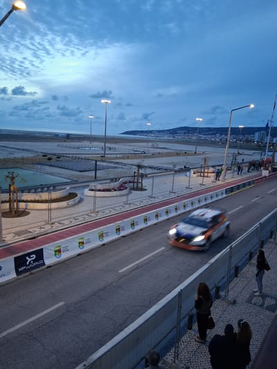 Captured from an elevated viewpoint, the image showcases a race car speeding along a road in Figueira da Foz, Portugal. Along the left side of the road, a beach is visible, adorned with streetlights that illuminate the scene. A pedestrian walkway borders the road, with people observing the race. The sky is a gradient of blues, suggesting either dawn or dusk.