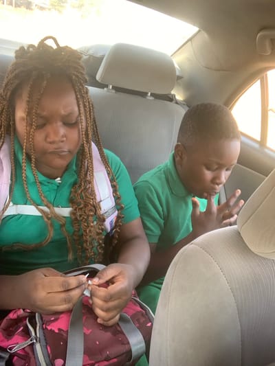 Two children are observed in the back seat of a vehicle in Kurudu, Nigeria. An older girl, with light-brown braided hair and a green polo shirt, wears a light-colored vest with white horizontal straps. She is seated on the left, looking downwards with a slight frown and pursed lips, manipulating a small, white object with both hands. A pink and maroon backpack with gray straps and metal zippers rests on her lap. To her right, a younger boy with short, dark hair wears a green polo shirt. He is looking downwards, with his fingers held near his mouth. The vehicle's interior features beige fabric seats and headrests. Daylight is visible through the windows on the right side, illuminating the scene from behind.