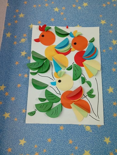 This media file showcases a colorful paper craft on a blue background adorned with yellow stars and a drawing of a ringed planet. The craft itself depicts a whimsical arrangement of circular and semi-circular paper cutouts in vibrant orange, red, green, yellow, and blue. These shapes are layered and folded to create stylized birds and foliage.
One prominent element is a yellow circle with a black dot for an eye, representing a bird's head. It has a red beak and red and green paper cutouts as a crest. This bird is surrounded by green, leaf-shaped cutouts that appear to be part of branches. Other birds are formed with orange circles for bodies, adorned with layered semi-circles of various colors for wings and tails. Some of these birds also have small red beaks and green leaf-like accents.
The arrangement suggests a playful, artistic creation, likely for educational or decorative purposes. The overall impression is cheerful and imaginative. The background, with its starry and planetary motifs, reinforces a theme of outer space or a fantasy world. No discernible text is visible in the image. The location context of Fergana City, Uzbekistan, is not visually indicated within the image itself.