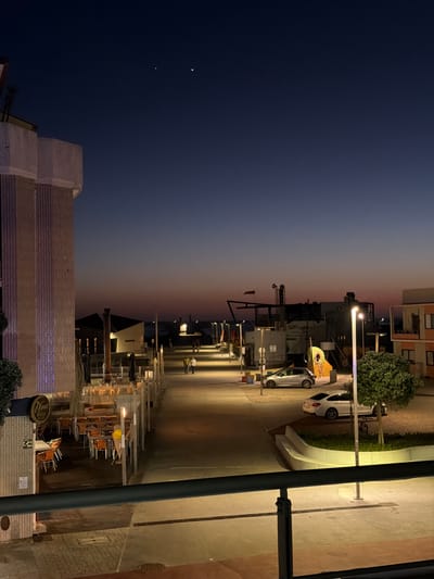 This detailed image depicts a twilight scene in Gafanha da Nazaré, Portugal, viewed from an elevated perspective, likely a balcony, indicated by a dark metal railing in the foreground. The sky transitions from a deep indigo overhead to a reddish-orange hue at the horizon. Two bright celestial objects are prominently visible in the upper-left quadrant of the sky.
On the left, a multi-story building with vertical textured panels is illuminated with a soft purple light. At its base, a restaurant named "Nova Barra" features outdoor dining tables with orange chairs under retracted umbrellas. A paved promenade extends from the foreground into the distance, brightly lit by modern streetlights with rectangular luminaires.
Several individuals are present on the promenade: two people are walking together in the mid-distance, and a solitary figure is also visible. Along the right side of the promenade, various low-rise buildings and commercial establishments are present. One building features a sign that reads "Caffé Lounge" and has an open-frame structure on its roof. Near it, a colorful, abstract playground structure or art installation is visible.
Two cars are parked on the right side of the promenade: a silver hatchback (identified as a Peugeot 208) and a white sedan (identified as a BMW 4-series Gran Coupe). A bicycle is parked near the white sedan. Maintained shrubbery is visible in planters, particularly a large bush in the right foreground adjacent to a streetlight. In the far distance, a faint horizon line suggests the presence of a body of water.