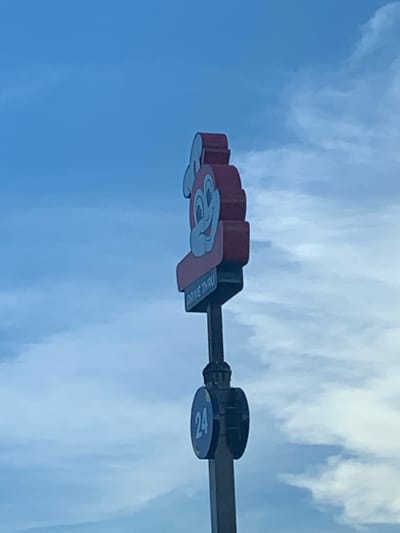 An outdoor, medium shot captures a sign for Jollibee's drive-thru in Angeles City, Philippines, against a backdrop of a hazy blue sky with scattered clouds. The Jollibee mascot, a stylized red bee with a wide smile and light blue accents, is prominently displayed at the top of the sign. Below the mascot, a sign with light blue text on a darker blue background reads "DRIVE THRU." Further down the pole, a circular sign with a dark blue background and yellow accents displays the number "24" in light blue. The pole itself is dark and appears to be metal. The image is taken from a low angle, looking up at the sign.