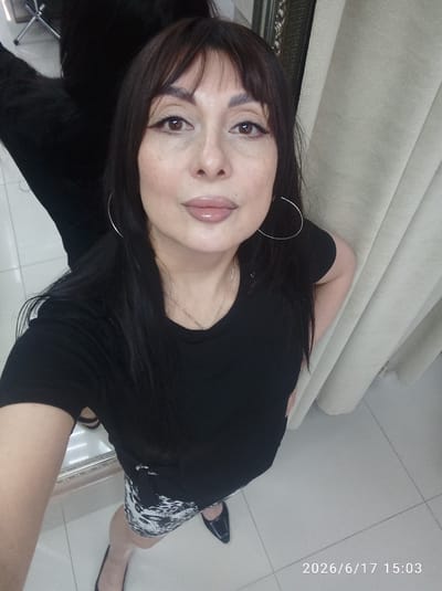 This is a medium shot, eye-level selfie of a woman, taken indoors. The woman has long, dark hair with bangs and is wearing a black t-shirt and a black and white patterned skirt. She has on hoop earrings and a necklace. Her makeup is applied with eyeliner and lip gloss. She is looking directly at the camera with a neutral expression.

The background consists of a beige curtain on the right and a large mirror reflecting a dark object, possibly a chair, on the left. The floor is tiled. The lighting appears to be artificial, suggesting it is daytime or evening.

The bottom right corner of the image displays the text "2026/6/17 15:03". There are no explicit indications of weather or specific events. The setting appears to be a room, possibly a dressing room or a bedroom.