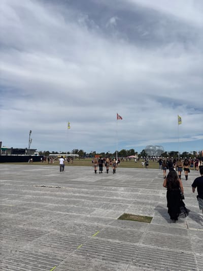 The image portrays an outdoor event occurring in Martínez, Argentina, under an overcast sky with visible cloud formations. The foreground consists of a large, grey, textured paved area, exhibiting a corrugated pattern and faint yellow linear markings. A square opening is present in the pavement in the mid-right.
Several individuals are depicted in various states of movement across the scene. In the immediate foreground, a person with long dark hair, wearing a dark, long dress or skirt, walks away from the camera towards the right. To their left, a group of four individuals, primarily women dressed in dark, short garments and boots, also walk away from the camera. Further left on the pavement, a male figure in a white shirt and dark pants walks away.
Beyond the paved section, a wide grassy field extends, populated by a significant crowd of people scattered across the area. On the far left, a substantial black stage structure with associated lighting towers is visible. Multiple tall flagpoles punctuate the field, displaying flags, including a red and white flag in the center-right and yellow flags further left and right. In the mid-ground on the right side, a large, translucent, multi-lobed inflatable structure stands. The horizon is lined with trees beneath the expansive cloudy sky.