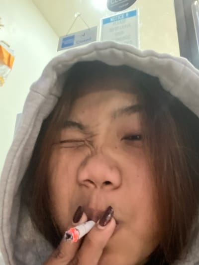 A close-up, low-angle image features an individual, likely female and of East or Southeast Asian descent, wearing a grey hooded sweatshirt with the hood up. She possesses long brown hair. Her right eye (viewer's left) is squinted or winked, and her lips are slightly puckered around a lit cigarette or rolled tobacco product held by her right hand. Her fingernails are long and painted a dark, possibly maroon or brown, color. The background reveals a light-colored wall where two signs are affixed. The left sign is blue and white, displaying the American Express logo and the word "Closed". The right sign is also blue and white, with "NOTICE !!" prominently displayed, followed by text advising against leaving valuables unattended, and the Indonesian word "TUTUP" (Closed) visible at the top. The scene is located within an indoor commercial setting in Uluwatu, Indonesia.
