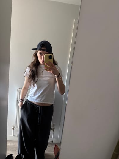 A young individual is depicted in a full-length mirror selfie taken indoors in Hull, United Kingdom. The person stands facing forward, wearing a dark blue baseball cap backward, a white cropped short-sleeved t-shirt revealing a portion of the abdomen, and dark gray loose-fitting sweatpants with a visible drawstring. A yellow smartphone is held in the left hand, pointed towards the mirror to capture the image, partially obscuring the lower face. The right hand is partially inserted into the right pocket of the sweatpants. A silver-colored chain necklace with a key-shaped pendant is visible, along with multiple bracelets on both wrists. The person's gaze is directed towards the camera with a slight pout. The background, reflected in the mirror, consists of a light-colored wall, a white panel radiator with associated piping, and light-colored wooden floorboards. Two dark objects, possibly shoes, are on the floor in the bottom left corner of the reflection, and a partial red object is visible on the floor in the bottom right.