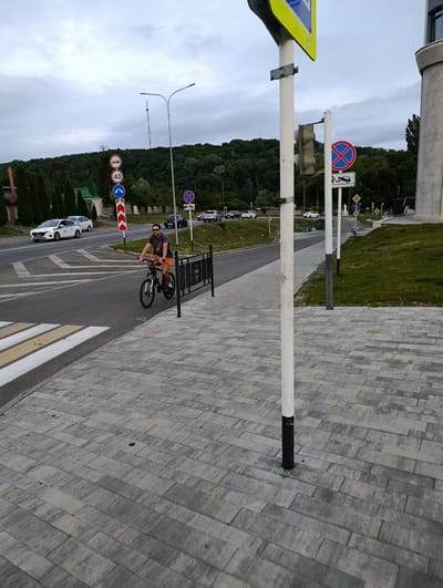 A man is riding a bicycle on a paved sidewalk next to a road. He is wearing a dark t-shirt, orange shorts, and sunglasses. The road has a pedestrian crossing with white and yellow stripes. Several cars are on the road, including a white police car. There are traffic signs, including a speed limit sign (40), a no stopping sign with a tow truck icon, and a left turn only sign. In the background, there are trees and a hill with a telecommunication tower. The sky is overcast. The location is identified as the city of Maykop, Russia.