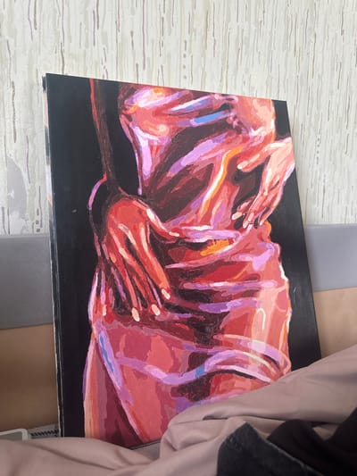The image shows a painting resting on a surface with a light-colored, wood-grain pattern. The painting features two figures in a reddish-purple color palette against a black background. The figures are intertwined, with hands appearing to reach out. Additional objects, including gray and brown textiles, are visible in the surrounding environment, along with sections of a beige fabric and part of a dark piece of clothing. The photograph's origin location is Высокий, Russia.