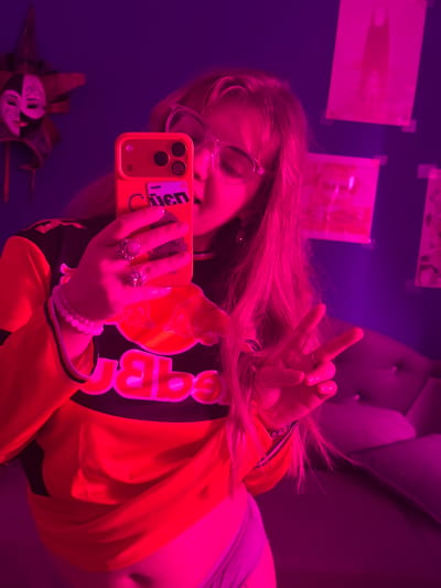 A person is taking a selfie in what appears to be a dimly lit room with a magenta/purple hue. The person is wearing glasses and an orange, black, and white jacket with the Red Bull logo prominently displayed. They are holding an orange iPhone with a sticker on the back. The person is making a peace sign with their fingers. There are rings on their fingers and a beaded bracelet on their wrist. A decorative mask is visible in the reflection below. On the wall are several papers or posters attached with tape. There is a tufted headboard visible in the background. I cannot confirm the specific location in Большая Шильна, Russia from this image.