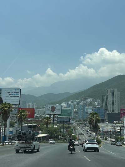 The image captures a daytime scene on a multi-lane highway in Monterrey, Mexico, looking towards a cityscape nestled against verdant, mountainous terrain. The sky is a pale blue with scattered, fluffy white clouds. In the foreground, a white pickup truck carrying a load of chairs is driving in the right lane. A motorcycle with a single rider is in the left lane, slightly ahead of the truck. A white Tesla sedan is in the far-right lane, further ahead. The highway is bordered by palm trees and various commercial advertisements, including a large AT&T billboard on the left promoting services in McAllen and Monterrey. The middle ground reveals a dense urban environment with numerous high-rise buildings of varying architectural styles and heights, some with glass facades. Residential buildings are scattered on the lower slopes of the mountains. The background is dominated by a large, green mountain range, partially obscured by haze. The overall impression is of a bustling metropolitan area integrated with its natural environment under a bright, partly cloudy sky.