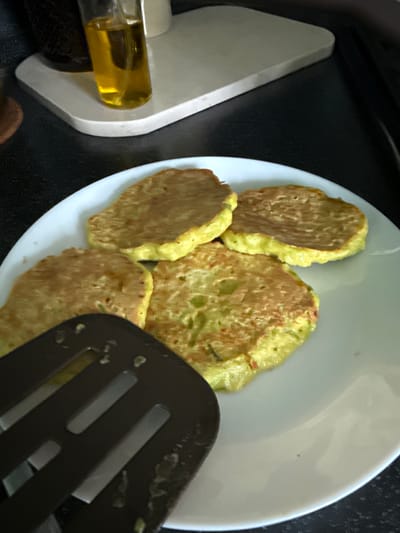 A close-up, slightly overhead shot shows a white bowl filled with several golden-brown, flat, circular fritters. The fritters appear to be made from a batter that includes finely chopped green vegetables, giving them a speckled appearance. One fritter in the foreground is partially obscured by a dark brown slotted spatula, which has some food residue on it. To the left of the bowl, a light gray, angled object with a clear glass container filled with yellow liquid is visible. The background is predominantly a textured black surface. The scene appears to be indoors, possibly in a kitchen, given the presence of cooking implements and the preparation of food. There is no visible text in the image.