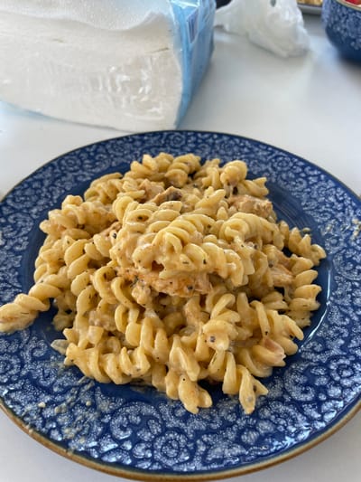A close-up shot shows a blue patterned plate filled with rotini pasta coated in a creamy sauce. Some small pieces of meat are visible within the pasta. To the left of the plate, a large stack of white paper napkins is partially visible, wrapped in clear plastic. The scene appears to be set on a white surface, possibly a table, with hints of a white tiled background. The lighting is bright and natural, suggesting an indoor setting during the day. The provided information about Skala Avdiron, Greece is not supported by the visual evidence in the image.