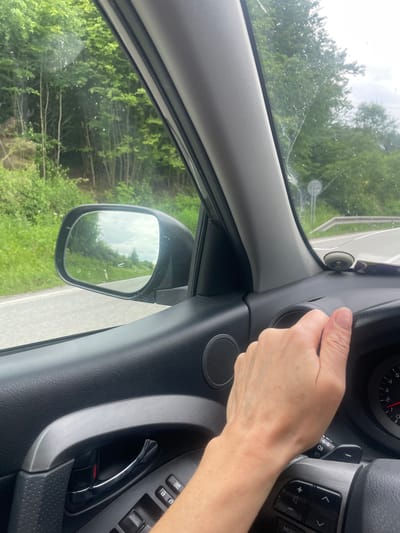 The image is taken from inside a car, presumably the driver's seat. A person's hand is visible near an air vent on the right side of the dashboard. Outside the car, through the driver's side window and side mirror, a road is visible with trees and greenery lining it. The windshield appears to have some damage, possibly cracks. The location is possibly Beňadikovce, Slovakia.