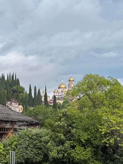 The image depicts a scenic view of New Athos, Abkhazia, featuring the iconic New Athos Monastery with its golden domes prominently displayed against a backdrop of lush green trees and a cloudy sky. In the foreground, the weathered roof of an old building, possibly a house or outbuilding, with a rusted satellite dish on its chimney, adds a touch of rustic charm. Tall cypress trees line the hillside, leading the eye towards the grand monastery situated on the elevation. The overall atmosphere is serene and picturesque, capturing the natural beauty and architectural heritage of the location.
