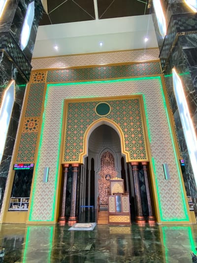 The image shows the interior of a mosque in Aluekambuk, Indonesia. The focal point is the richly decorated mihrab, the niche indicating the direction of prayer, which is adorned with intricate geometric patterns in gold, green, and orange. The mihrab is framed by tall, dark marble columns with copper accents. Green LED lights illuminate the decorative borders of the mihrab and the surrounding walls, which are also covered in a patterned wallpaper. In front of the mihrab, a wooden minbar (pulpit) stands, with a Quran placed on its top. A prayer mat is laid out on the floor in front of the minbar. The floor itself is made of polished dark green marble, reflecting the surrounding lights. On the left side of the image, a digital display shows time and prayer schedules. The overall impression is one of grandeur and ornate Islamic architecture. There are no people visible in the image.