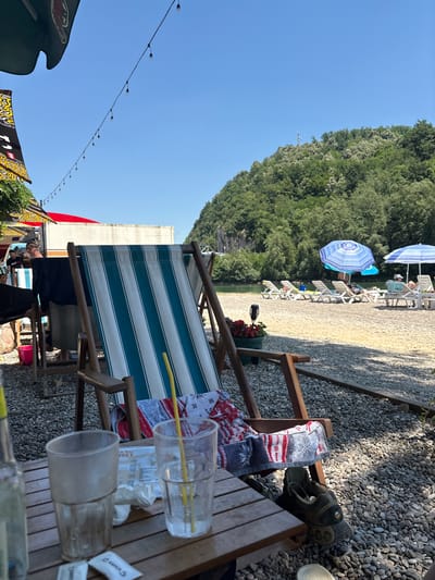 This image depicts a sunny outdoor seating area at a beach bar, likely located in Zvornik, Bosnia and Herzegovina. In the foreground, a wooden table holds two empty drinking glasses, one with a yellow straw. A bottle is also partially visible on the left side of the table. A striped deck chair, featuring teal and white stripes, is prominent in the center, draped with a towel. Adjacent to the chair, a pair of hiking shoes rests on the gravelly ground. Behind the chair, several other deck chairs are set up on a sandy and pebbly beach, some occupied by individuals relaxing under blue and white umbrellas. The background reveals a lush, tree-covered hill sloping down towards a body of water. The sky is clear and blue. String lights are visible overhead, strung between unseen structures, suggesting an evening ambiance.