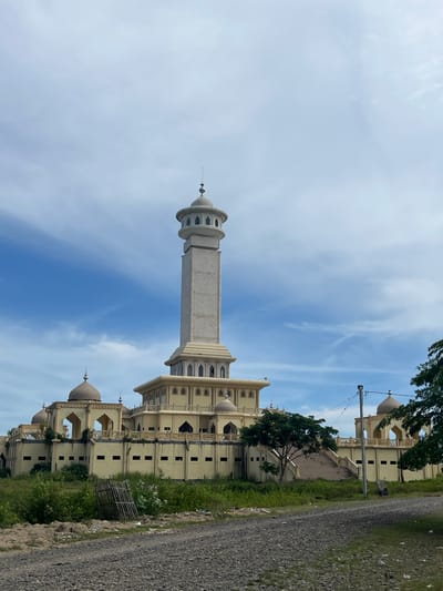 An exterior, eye-level, wide shot captures a large, pale yellow mosque with a tall, slender minaret against a bright blue sky with scattered white clouds. The mosque features multiple domes and arched walkways, with a prominent central tower topped by a smaller dome. The building is surrounded by lush green vegetation, including trees and overgrown grass. In the foreground, a gravel road leads towards the structure, with a broken wooden fence on the left and some debris on the right. The scene appears to be located in Kruengmate, Indonesia, as indicated by the distinct architectural style and the surrounding tropical foliage. There are no people or animals visible in the image. The overall impression is one of a grand but somewhat isolated and possibly unfinished religious complex.