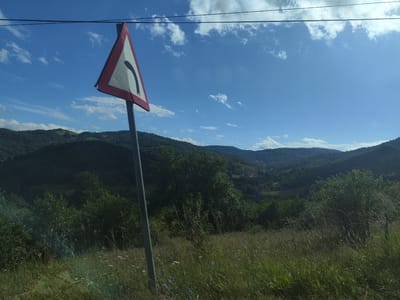 The image depicts a scenic view of rolling, tree-covered hills under a bright blue sky with scattered white clouds. In the foreground, a traffic sign warning of a sharp left turn is prominently displayed on a metal pole. The sign is triangular with a red border and a white interior, featuring a black curved arrow indicating the turn. Surrounding the sign is a field of tall, green grass with some wildflowers. Power lines are visible in the upper portion of the image, stretching across the sky. The landscape suggests a rural or mountainous area, consistent with locations like Petrova, Romania, known for its natural beauty and hilly terrain. There are no people visible in the image. The overall atmosphere is serene and natural.