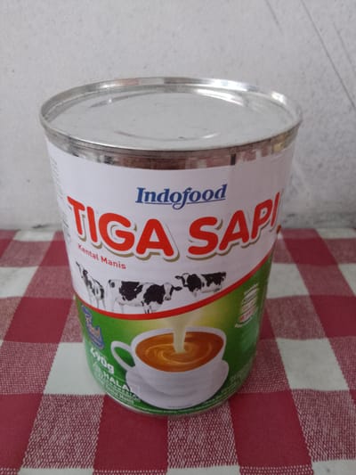 A close-up, eye-level, medium shot captures a cylindrical metal can of Indofood Tiga Sapi sweetened condensed milk, prominently displayed on a red and white checkered tablecloth. The can's label is predominantly white with bold red lettering that spells out "TIGA SAPI," with "Indofood" in blue above it. Below the brand name, "Kental Manis" is written in smaller black text. A stylized illustration of cows is featured across the middle of the can. The lower portion of the label shows a white cup with a brown beverage being poured into it, and a green band encircling the bottom. Text in various sizes and colors, including "490g" and "HALAL," is visible on the lower section of the can. The background consists of a textured, light-colored wall, suggesting an indoor setting. The lighting is neutral, with no strong shadows or highlights. The image composition emphasizes the product packaging. This scene is situated in Cisarua, Indonesia.