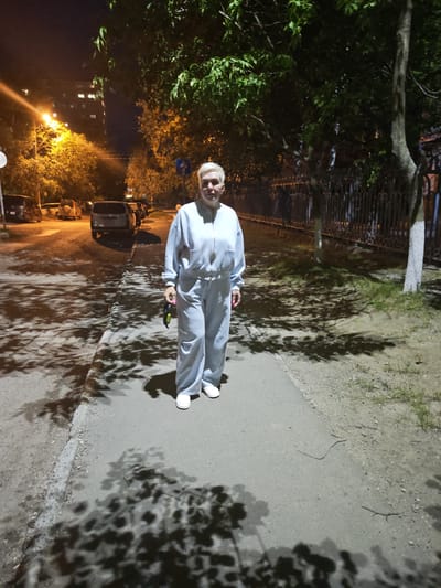 A woman with short, blonde hair is standing on a sidewalk at night. She is wearing a light blue tracksuit and white sneakers. In her right hand, she is holding a dark object with a yellow end. She is facing the camera with a neutral expression. The sidewalk is next to a street with parked cars. Trees with green leaves cast shadows on the sidewalk and street. A metal fence runs along the side of the sidewalk, beyond which is grass and more trees. In the background, there is a building with illuminated windows and streetlights. The scene appears to be a residential street in the city of Ussurijsk, Russia.