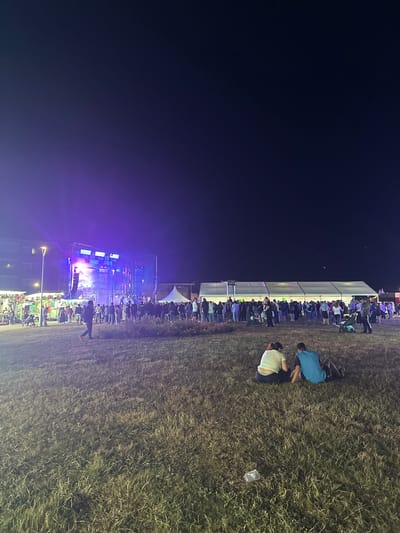 The image depicts an outdoor evening event at El Muséu del Pueblu d'Asturies in Gijón, Spain. In the foreground, two individuals are seated on the dry grass, facing away from the camera. The middle ground shows a crowd of people gathered in front of a large stage with bright purple lights. To the right, a long white tent stands, also with a gathering of people in front of it. The background features buildings and a dark, starry sky. The overall atmosphere suggests a festival or concert setting.