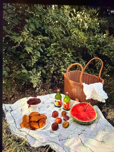 The image displays an overhead view of a picturesque picnic spread laid out on a white blanket with green embroidered details, set amidst lush green foliage. The scene suggests an outdoor gathering in a natural, possibly verdant park or garden setting, consistent with the location context of Ros Na Rí, Ireland. No people are visible.

A prominent wicker picnic basket, containing a ruffled white cloth, sits on the left side of the blanket. Adjacent to it, a large, half-cut watermelon, vibrant red, has a wooden skewer or utensil inserted into its flesh. Nearby, two green glass bottles are visible, both clearly labeled with the brand name "Yummal!". Several dark red and purple plums or nectarines are scattered on the blanket, some appearing halved.

Towards the bottom right of the blanket, a white bowl is filled with multiple golden-brown croissants, and a smaller white cup or bowl contains a generous portion of small red berries, possibly cranberries or currants.

The surrounding environment is dominated by dense green bushes and leafy plants, some adorned with tiny white blossoms, indicating a rich natural backdrop. The lighting appears soft and natural, suggesting a pleasant daytime, possibly late afternoon, suitable for outdoor dining. The overall atmosphere conveys a sense of tranquility and enjoyment of nature with an inviting meal. In the upper left corner of the original image, the text "1/1" is faintly visible.