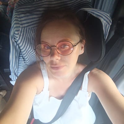 A close-up, high-angle selfie shows a woman in a car. She is wearing round, pink-tinted glasses, a white tank top, and a blue and white striped shirt. A black seatbelt is visible across her chest. The background consists of the car's interior, including a headrest and the seatbelt mechanism. The image is taken indoors, likely during the day. There is no indication of the city of Karacha-Elga, Russia.