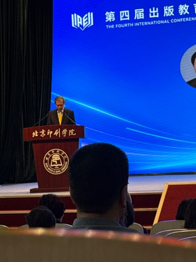 The image captures a scene from an indoor conference or event, likely at the Beijing Institute of Graphic Communication, given the text on the podium. A Caucasian man in a suit and yellow tie stands at a podium, looking down and speaking. The podium is dark red with gold lettering that reads "北京印刷学院" (Beijing Institute of Graphic Communication). Behind the speaker, a large blue screen displays Chinese characters and English text: "第四届出版教育国际研讨会 THE FOURTH INTERNATIONAL CONFERENCE". A circular logo is also visible on the podium. In the foreground, the backs of several attendees' heads are visible, with one person's head in sharp focus, obscuring the lower portion of the podium. The environment appears to be a lecture hall or auditorium with dark curtains on the left side. The setting is consistent with an event held in Daxing District, China, where the Beijing Institute of Graphic Communication is located.